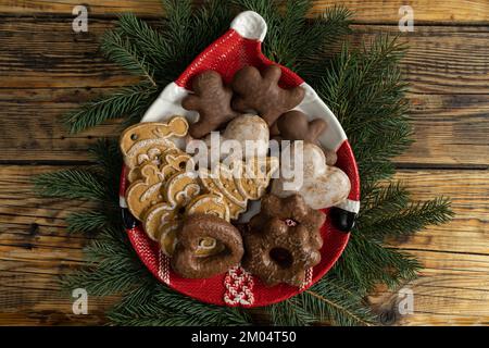 Lebkuchen-Weihnachtskekse auf einem Santa-förmigen Teller und Ästen. Weihnachtszeit Winterkomposition auf Holzhintergrund. Stockfoto