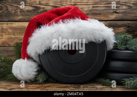 Gym Kurzhantel Hantelscheibe in rotem Weihnachtsmann Hut und Weihnachtsbaumzweigen. Gesunde Fitness-Feriensaison, Winter-Workout Zusammensetzung. Stockfoto