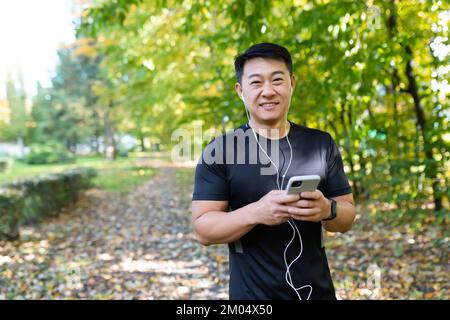 Porträt eines jungen, gutaussehenden Asiaten, der morgens im Park joggen ging. Er hält ein Telefon in der Hand, hört Musik über Kopfhörer, sieht in die Kamera, lächelt Stockfoto