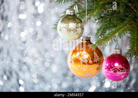 Weihnachtsdekorationen. Drei klassische Weihnachtsbälle in verschiedenen Farben mit Zeichnungen auf dem Weihnachtsbaum und Schneeflocken auf einem silbernen Bokeh Backgro Stockfoto