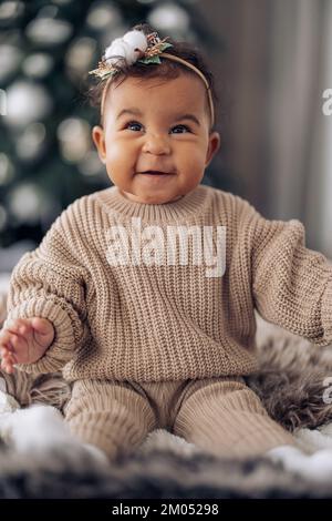 Ein glückliches Baby mit gemischter Rasse sitzt auf dem Bett vor dem Hintergrund des Weihnachtsbaums. Der Begriff der zwischenrassischen Familie und die Einheit zwischen verschiedenen menschlichen Rassen. Stockfoto