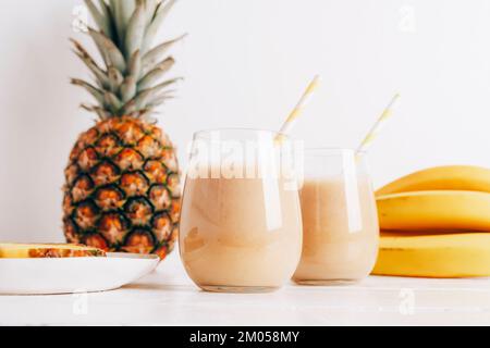 Stillleben mit Obst, Banane, Ananas und Smoothie in Gläsern auf einem weißen Holztisch. Minimal-Entgiftungsdiät-Konzept, Sommer-Vitamingetränk. Vorderansicht. Stockfoto