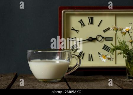 eine tasse Milch und eine Vase mit Gänseblümchen in der Nähe der Uhr auf einem Holztisch Stockfoto