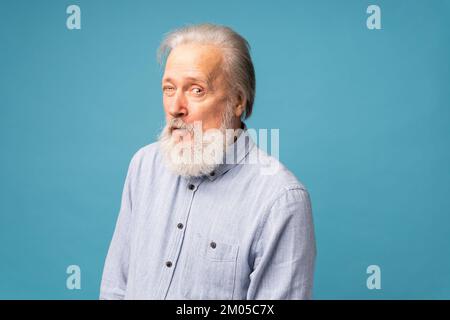 Porträt eines weißen bärtigen, ehrlichen alten Mannes Wink Tragen Sie gut aussehende Kleidung isoliert auf blauem Hintergrund - positive menschliche Emotionen Gesichtsausdruck Stockfoto