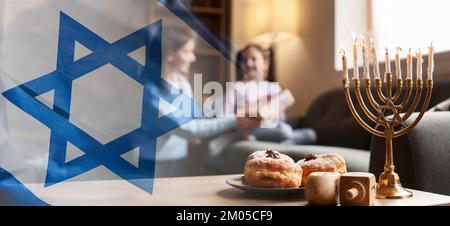 Banner mit israelischer Nationalflagge und Menorah, Dreidel und Donuts auf dem Tisch einer glücklichen Familie, die Hannukah zu Hause feiert Stockfoto