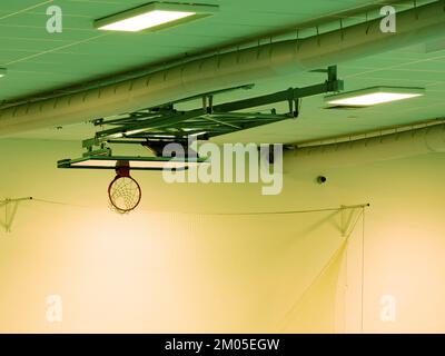 Gefalteter Basketballkorb mit Balg an der Decke. Der Korb mit weißem Netz im Innensportdach mit Metallhintergrund Stockfoto