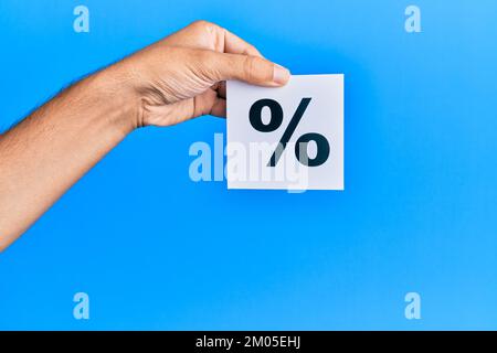 Hand eines kaukasischen Mannes, der Papier mit einer Prozentmarke auf einem isolierten blauen Hintergrund hält Stockfoto