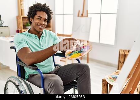 Junger afroamerikanischer behinderter Künstlermann, der im Kunststudio auf dem Rollstuhl sitzt und zeichnet. Stockfoto