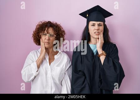 Hispanische Mutter und Tochter, die eine Abschlussmütze und einen Morgenmantel tragen und den Mund mit der Hand berühren, mit schmerzhaftem Gesichtsausdruck wegen Zahnschmerzen oder Zahnschmerzen Stockfoto