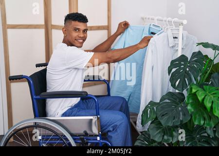 Ein junger lateiner, der ein Hemd aussucht, sitzt im Rollstuhl im Schlafzimmer Stockfoto
