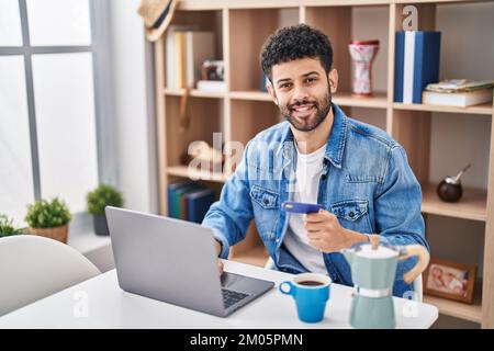 Junger arabischer Mann mit Laptop und Kreditkarte, der zu Hause auf dem Tisch sitzt Stockfoto