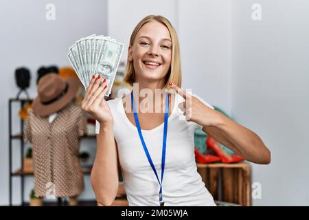 Junge weiße Frau, die in einer Boutique arbeitet und Dollarscheine in der Hand hält, die fröhlich mit Hand und Finger zeigen Stockfoto