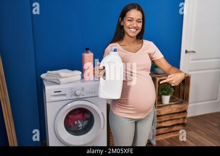 Junge schwangere Frau, die Wäsche wäscht und eine Waschmittelflasche hält, die mit dem Finger auf ein selbstlächelndes, glückliches und stolzes Gesicht zeigt Stockfoto