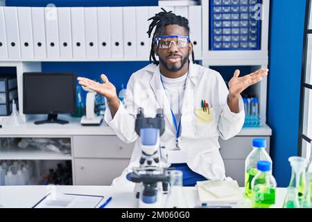 Afrikanischer Mann mit Dreadlocks, der im Labor arbeitet, schreit und schreit laut zur Seite, mit der Hand auf dem Mund. Kommunikationskonzept. Stockfoto