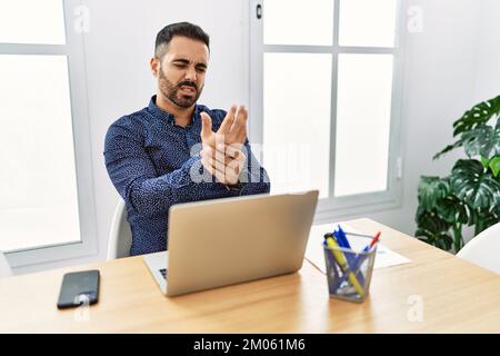 Junger hispanischer Mann mit Bart, der im Büro arbeitet, mit einem Laptop, der Schmerzen an Händen und Fingern hat, Arthritis, Entzündung Stockfoto