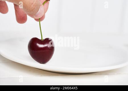 Nahaufnahme einer Frauenhand, die eine herzförmige Kirsche auf einem weißen Teller hält. Reife, frische, saftige Beere auf weißem Hintergrund. Mit Platz zum Kopieren. Hochwertiges Foto Stockfoto