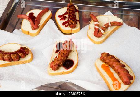 Hot Dog mit verschiedenen Gewürzen und Chili, Paprika und Tomaten und Soßen, Senf und Ketchup auf einem eisernen Tablett auf einem weißen Tisch. Stockfoto