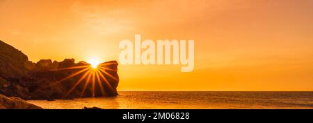 Banner. Ein epischer Sonnenuntergang auf dem Meer - die Sonne geht hinter dem Berg unter und alles scheint mit warmem Licht und langen Strahlen. Stockfoto