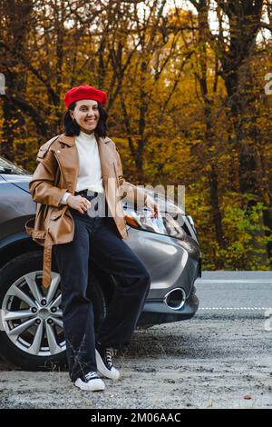 Eine junge Frau in einer roten Baskenmütze steht in der Nähe des Wagens vor dem Hintergrund eines Herbstwaldes Stockfoto