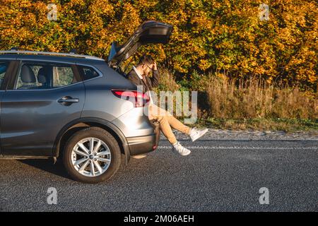 In der Nähe des Herbstwaldes sitzt ein Mann im Kofferraum seines Autos und ruht sich nach einer langen Reise aus Stockfoto