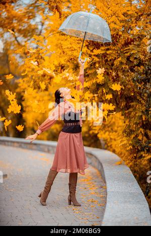 Hübsches Mädchen in einem Kleid mit Schirm im Herbstpark. Sie hält ihn über den Kopf, die Herbstblätter fallen aus ihm heraus Stockfoto