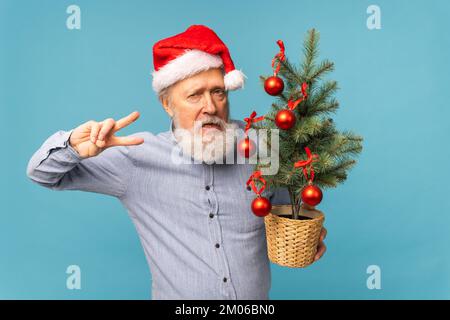 Portrait von fröhlichen, verrückten Emotionen der Weihnachtsmann schaute in die Kamera und hielt einen kleinen weihnachtsbaum auf blauem Hintergrund Stockfoto