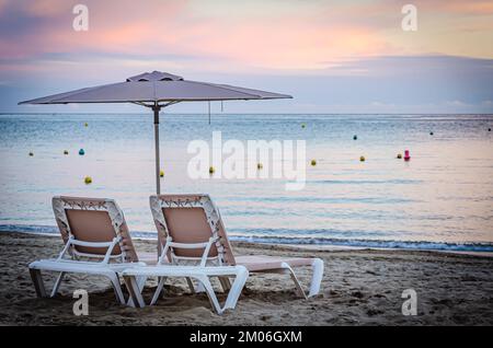 Zwei Sonnenliegen und Sonnenschirme am Strand mit pinkfarbenem Sonnenuntergang im Hintergrund Stockfoto