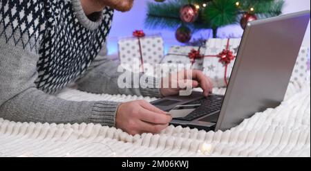 Mann mit Laptop und Kreditkarte kauft Geschenke für Neujahr 2023, Weihnachten. Ein Mann im Weihnachtsmütze auf dem Hintergrund von Weihnachtsbaum, Girlanden, Geschenken mit einem Laptop. Cybersicherheit vor Betrügern. Neon-Bokeh. Stockfoto