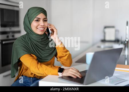 Muslimische Frau im Hidschab, die mit dem Handy spricht und in der Küche ein Notebook benutzt Stockfoto