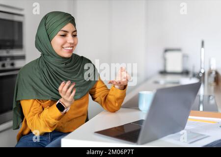 Telekonferenz. Lächelnde Junge Muslimische Frau, Die Videoanrufe Mit Einem Laptop In Der Küche Macht Stockfoto