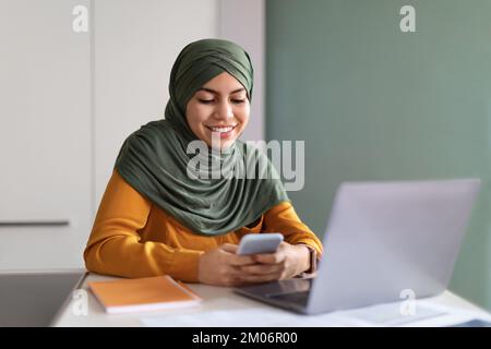 Lächelnde Junge Muslimische Frau In Hijab Mit Smartphone Im Büro Stockfoto