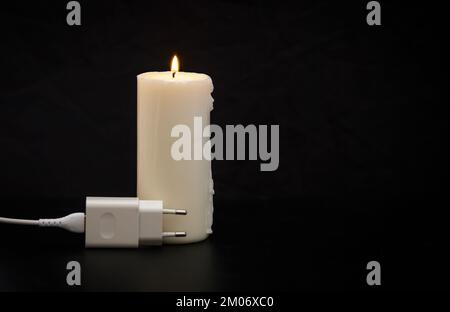 Blackout-Konzept. Ladegerät mit Kabel in der Nähe der Kerze auf dunklem Hintergrund. Stockfoto