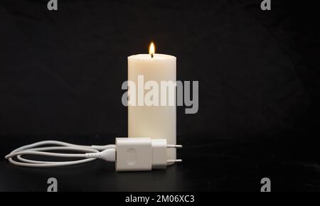 Blackout-Konzept. Ladegerät mit Kabel in der Nähe der Kerze auf dunklem Hintergrund. Stockfoto