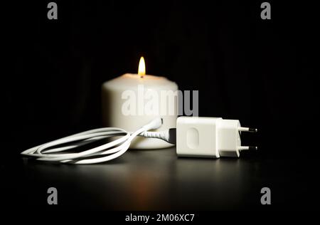 Blackout-Konzept. Ladegerät mit Kabel in der Nähe der Kerze auf dunklem Hintergrund. Stockfoto