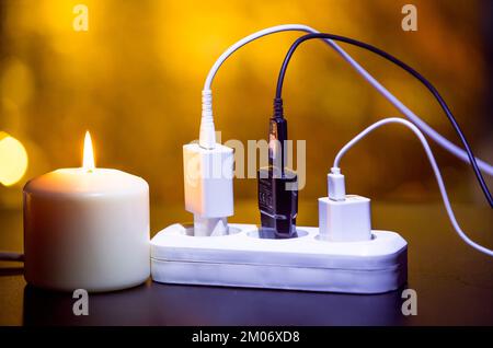 Blackout-Konzept. Ladegerät mit Kabel in der Nähe der Kerze auf dunklem Hintergrund. Stockfoto