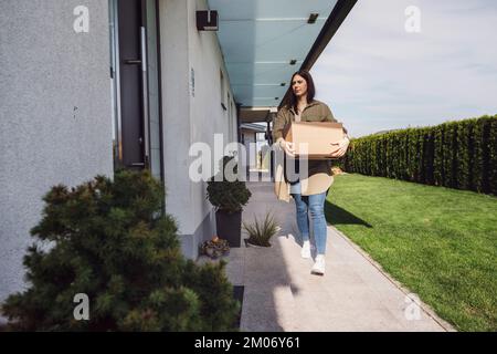 Junge weiße Frau, die eine Schachtel trug, die sie von der Post bis zur Haustür ihres Hauses bekam Stockfoto