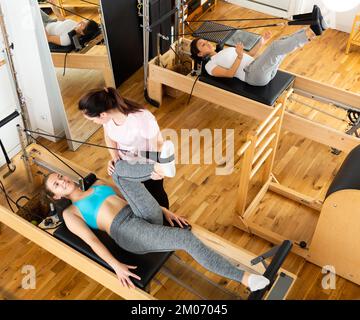 Blick von oben auf die Trainerin, die jungen Frauen beim Pilates-Üben auf Reformer hilft Stockfoto