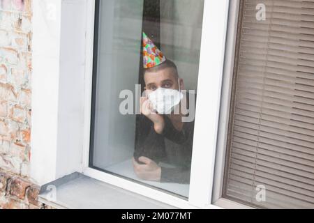 Aufgewühlter junger Mann, der an seinem Geburtstag aus dem Fenster schaut. Das Konzept der Einsamkeit in der Quarantäne während der Coronavirus-COV-Pandemie Stockfoto