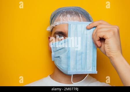 Der Kerl zieht eine Atemschutzmaske über seine Augen. Ein attraktiver Mann setzt eine Maske auf und schaut in die Kamera. Erkältungen, Grippe, Virus, Tonsillitis, akute Atmung Stockfoto