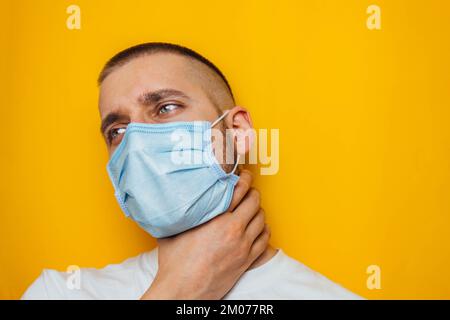 Der Typ hält seine Kehle mit der Hand und zeigt, dass es weh tut. Der maskierte Mann schaut in die Kamera. Erkältungen, Grippe, Virus, Quarantäne, Epidemie Stockfoto