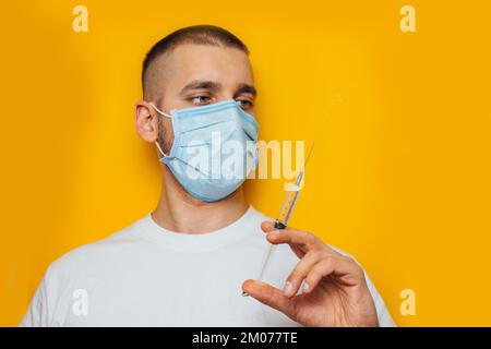 Porträt eines jungen Mannes in einem Atemgerät auf gelbem Hintergrund. Halten einer Ampulle mit einem Coronavirus-Impfstoff. Erkältungsmedizin. Das Konzept des Corerivas Stockfoto