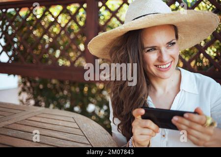 Glückliche junge Frau in weißem Hemd mit Hut, die am Tisch sitzt und SMS mit Smartphone auf der Terrasse des Gästehauses sendet. Stockfoto