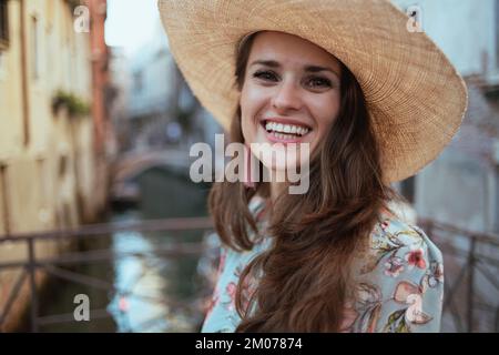 Portrait einer glücklichen, modernen allein Reisenden Frau in Blumenkleid mit Hut, die die Promenade in Venedig, Italien, genießt. Stockfoto