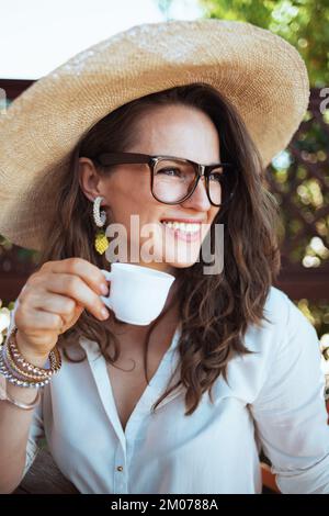 Lächelnde Hausfrau mittleren Alters in weißem Hemd mit einer Tasse Kaffee, Hut und Brille auf der Terrasse. Stockfoto