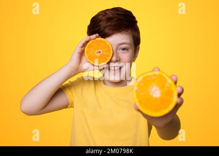 Der Junge hält zwei orangefarbene Scheiben in den Händen, bedeckt sein Auge mit einer und hält die andere gegen die Kamera isoliert auf gelbem Hintergrund. Stockfoto