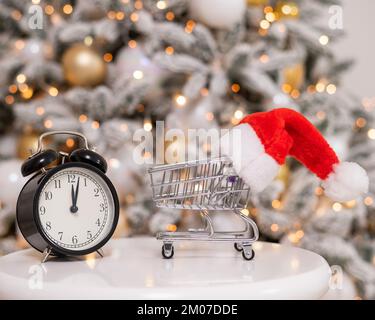 Weihnachtsgeschenke Zeit zum Einkaufen. Ein Einkaufswagen mit Weihnachtsmütze und Wecker am Weihnachtsbaum. Stockfoto