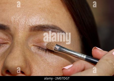 Make-up-Künstler wendet Lidschatten an . Ein wunderschönes Frauengesicht. Hand der Visagiste, Malkosmetik des jungen Schönheitsmodellmädchens. Nachholprozess läuft Stockfoto