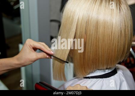 Frisur. Eine attraktive Blondine mit kurzen Haaren macht eine Frisur mit einem elektrischen Bügeleisen. Grauer Hintergrund des Haarglätters. Nahaufnahme. Makrofoto Stockfoto