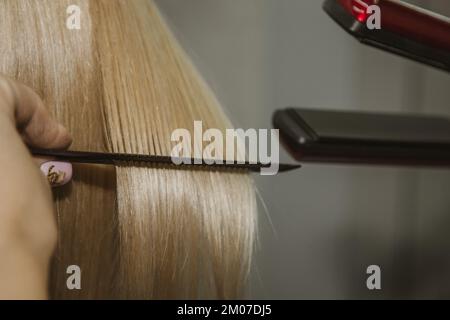 Frisur. Eine attraktive Blondine mit kurzen Haaren macht eine Frisur mit einem elektrischen Bügeleisen. Grauer Hintergrund des Haarglätters. Nahaufnahme. Makrofoto Stockfoto