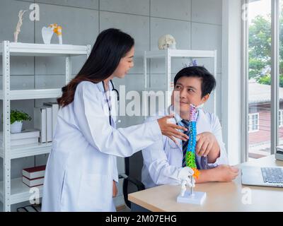 Zwei asiatische Ärzte in weißen Uniformen unterhalten sich und arbeiten im medizinischen Büro zusammen. Der orthopädische Erwachsene männliche Arzt vermittelt Wissen und Beratung Stockfoto